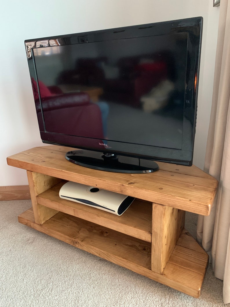 Puede incluir: Un mueble de TV r&uacute;stico de madera con un televisor de pantalla plana negro. El mueble tiene un acabado de madera natural y un dise&ntilde;o de esquina. Una consola de juegos blanca se encuentra en el estante del medio. El televisor es de la marca Technika.