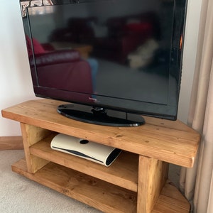 Puede incluir: Un televisor de pantalla plana negro se encuentra sobre un mueble de esquina de madera r&uacute;stica de color marr&oacute;n claro. El mueble tiene dos estantes y un acabado de vetas de madera natural. Un dispositivo electr&oacute;nico blanco est&aacute; en el estante del medio.