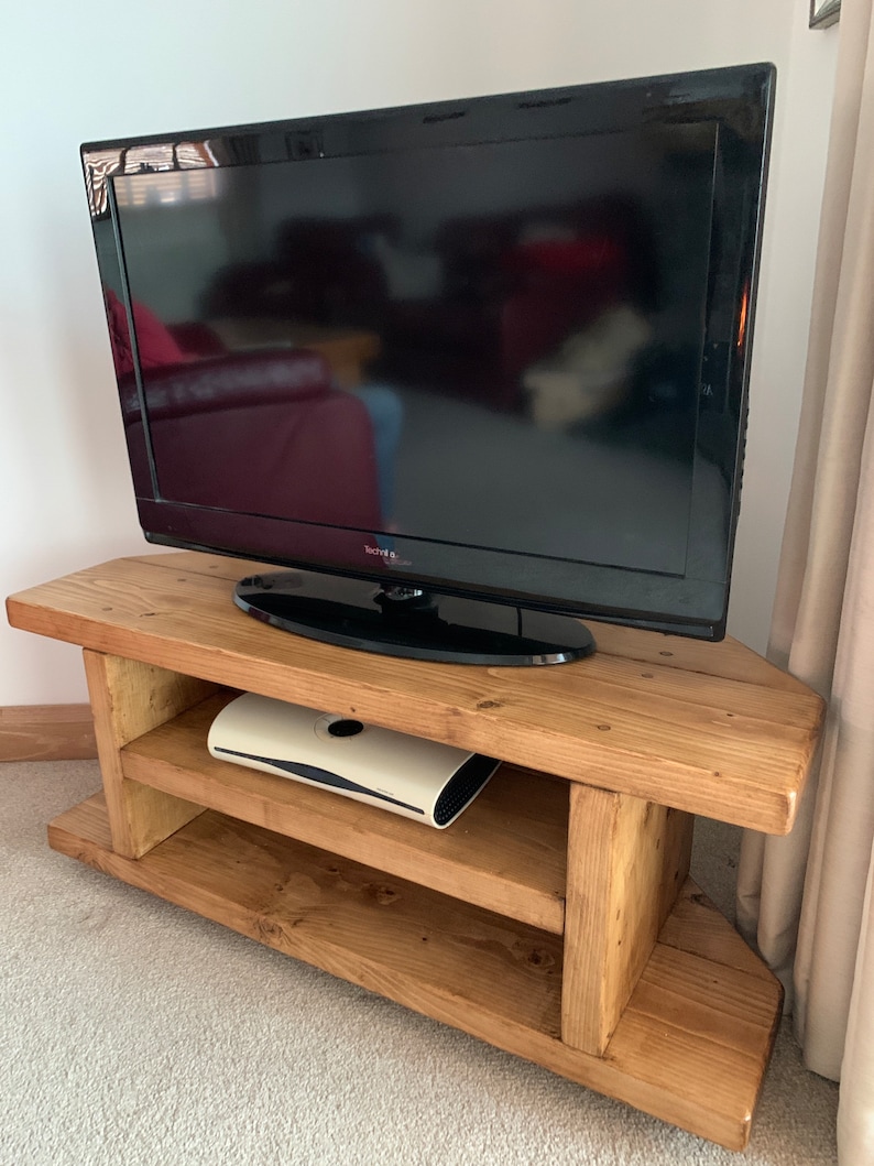 Puede incluir: Un televisor de pantalla plana negro se encuentra sobre un mueble de esquina de madera r&uacute;stica de color marr&oacute;n claro. El mueble tiene dos estantes y un acabado de vetas de madera natural. Un dispositivo electr&oacute;nico blanco est&aacute; en el estante del medio.