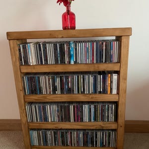 May include: A wooden CD storage unit filled with compact discs. A red glass vase with a red flower arrangement sits on top. The unit has four shelves and is made of light brown wood.