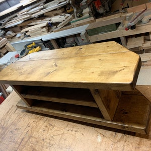 Puede incluir: Un zapatero de madera hecho a mano con un acabado natural. El zapatero tiene una parte superior rectangular con esquinas redondeadas, dos estantes y una base. La veta de la madera es visible. La pieza es de madera maciza.