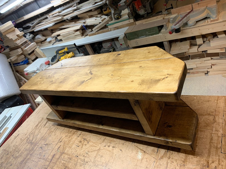 Puede incluir: Un zapatero de madera hecho a mano con un acabado natural. El zapatero tiene una parte superior rectangular con esquinas redondeadas, dos estantes y una base. La veta de la madera es visible. La pieza es de madera maciza.