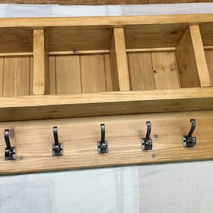 May include: A wooden wall shelf unit with five storage compartments and six metal hooks. The light-coloured wood unit is designed for wall mounting, ideal for an entryway or hallway.