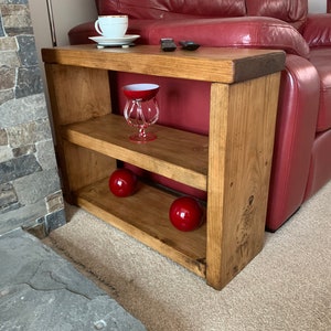 May include: A wooden side table with two shelves, stained a warm brown. A white teacup, two black stones, a red glass, and two red spheres are displayed on the table. The table is placed next to a red leather sofa.