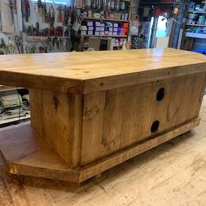 Puede incluir: Consola de medios de madera hecha a mano con un acabado natural. La consola presenta una parte superior rectangular con bordes redondeados, un estante inferior y dos aberturas circulares en la parte delantera. La veta de la madera es visible y la pieza est&aacute; sobre peque&ntilde;os pies.