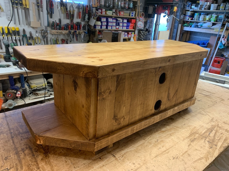 Puede incluir: Consola de medios de madera hecha a mano con un acabado natural. La consola presenta una parte superior rectangular con bordes redondeados, un estante inferior y dos aberturas circulares en la parte delantera. La veta de la madera es visible y la pieza est&aacute; sobre peque&ntilde;os pies.