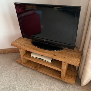 Puede incluir: Un televisor Toshiba negro se encuentra sobre un mueble de TV r&uacute;stico de madera marr&oacute;n claro. El mueble tiene dos estantes y un dise&ntilde;o en &aacute;ngulo. El mueble de TV est&aacute; sobre una alfombra gris claro.