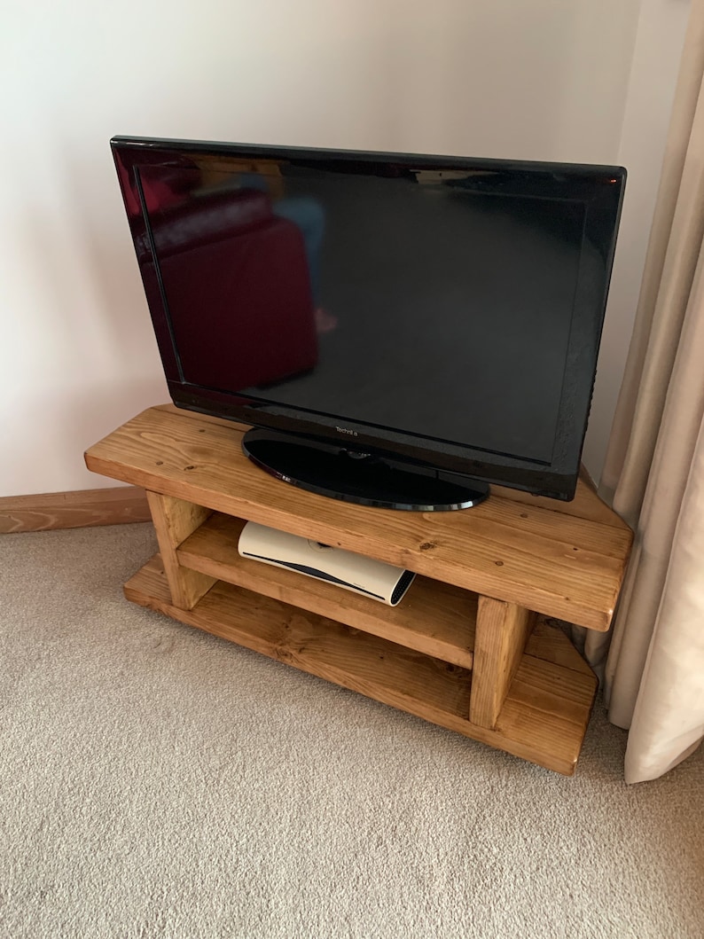Puede incluir: Un televisor Toshiba negro se encuentra sobre un mueble de TV r&uacute;stico de madera marr&oacute;n claro. El mueble tiene dos estantes y un dise&ntilde;o en &aacute;ngulo. El mueble de TV est&aacute; sobre una alfombra gris claro.