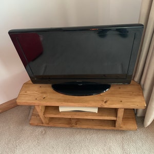 Puede incluir: Un televisor Sony negro se encuentra sobre un mueble de TV r&uacute;stico de madera marr&oacute;n claro. El mueble tiene dos estantes y est&aacute; colocado en una esquina de una habitaci&oacute;n con alfombra beige y una pared blanca.
