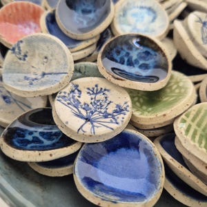 May include: A selection of small, handmade ceramic dishes in shades of blue, white, and green. Some have floral or patterned designs. The dishes have a rustic, artisanal appearance, with visible texture and colour variations.