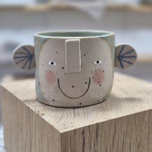 Peut inclure: Une tasse en céramique blanche avec une anse noire et le texte "But First, Coffee" en noir, dans une police manuscrite. La tasse est posée sur une surface blanche.