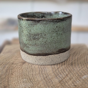 Peut inclure: Un gobelet en céramique fait à la main avec un glaçage vert d'eau moucheté. Le bord supérieur et l'intérieur ont une teinte marron plus foncée, tandis que la base est beige naturel non émaillé. La tasse cylindrique repose sur une surface en bois.