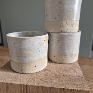 Peut inclure: Quatre tasses en céramique faites à la main. Les tasses sont cylindriques avec une moitié supérieure gris clair moucheté et une base de couleur crème. Elles sont empilées sur un bloc de bois, mettant en valeur leur design rustique et artisanal. Les tasses sont idéales pour le café ou le thé.