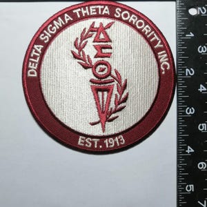May include: Circular embroidered patch with a maroon border and a light gray center. The text "DELTA SIGMA THETA SORORITY INC." is in maroon around the edge. The center features a maroon torch and the text "EST. 1913".