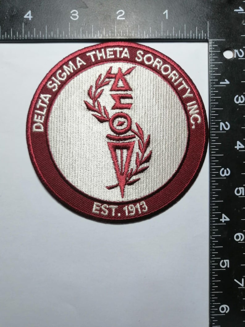 May include: Circular embroidered patch with a maroon border and a light gray center. The text "DELTA SIGMA THETA SORORITY INC." is in maroon around the edge. The center features a maroon torch and the text "EST. 1913".