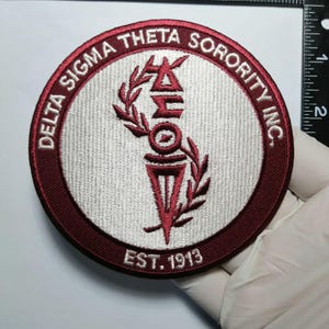 May include: A round, embroidered patch with the text "DELTA SIGMA THETA SORORITY INC." in burgundy. The center features the sorority's symbols in burgundy and white, with "EST. 1913" below. The patch is approximately 3 inches in diameter.
