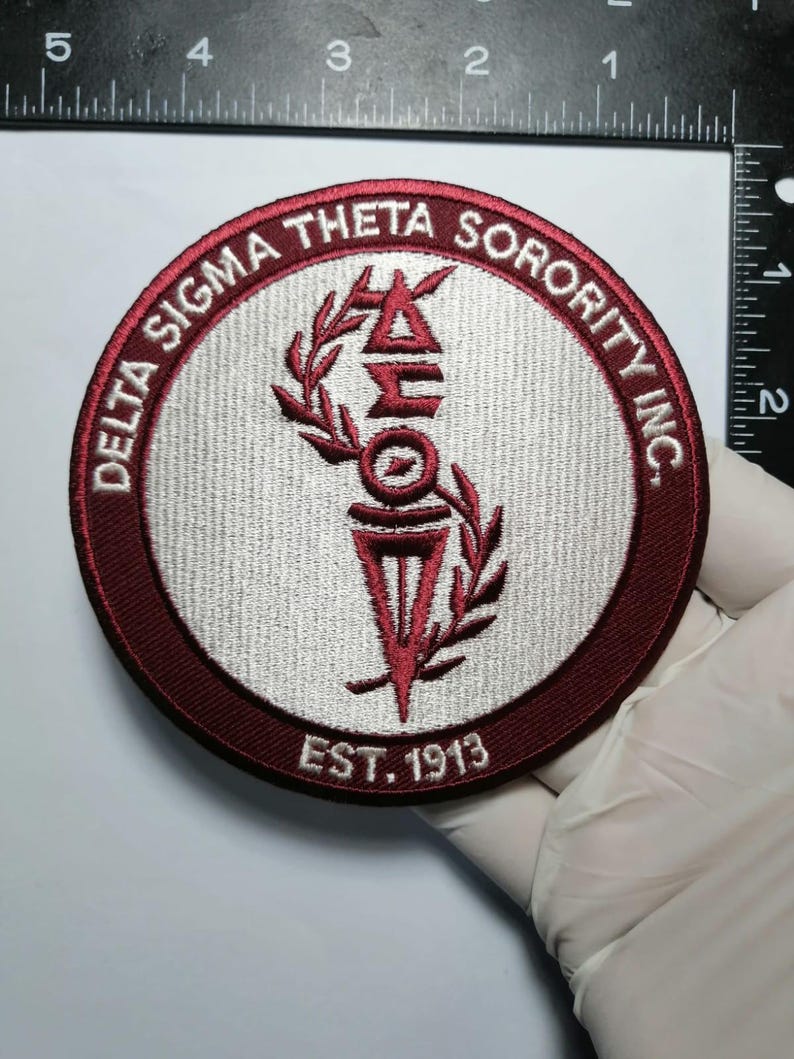 May include: A round, embroidered patch with the text "DELTA SIGMA THETA SORORITY INC." in burgundy. The center features the sorority's symbols in burgundy and white, with "EST. 1913" below. The patch is approximately 3 inches in diameter.