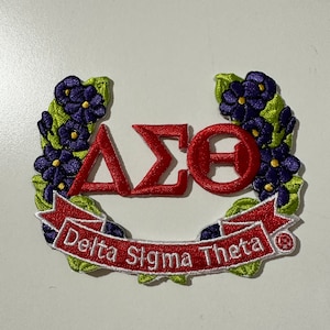 May include: Embroidered patch featuring the Greek letters Delta, Sigma, and Theta in red, surrounded by a floral wreath with purple flowers and green leaves. A red banner displays the text "Delta Sigma Theta" in white.
