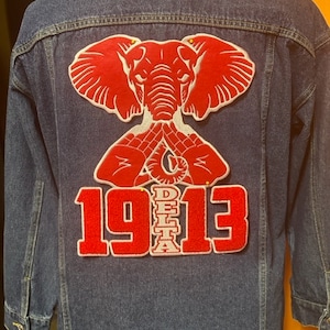 May include: A denim jacket with a large red elephant and "1913" patch on the back. The elephant design is detailed with white outlines. The jacket is a medium wash blue.