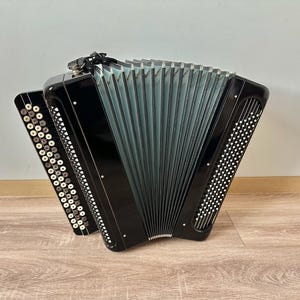 Könnte beinhalten: Ein schwarzes Akkordeon mit einem silbernen und weißen Knopfklangfeld. Das Akkordeon ist geschlossen und hat einen schwarzen Lederriemen.