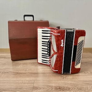 Op de afbeelding: Een rode accordeon met witte toetsen en zwarte accenten, naast een bruine draagkoffer. De accordeon heeft de naam "Maia" erop. De instrumenten staan op een houten vloer.