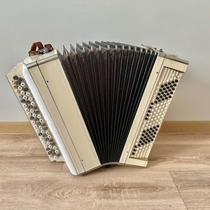 May include: An antique accordion with a cream-colored body and black bellows. The instrument features rows of buttons and a brown leather handle. The accordion is positioned on a wooden floor against a neutral wall.