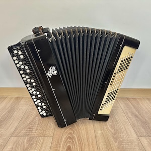 Könnte beinhalten: Ein schwarzes Knopfakkordeon mit einem zentralen Balg, einer Tastatur mit zahlreichen weißen und schwarzen runden Knöpfen sowie einem dekorativen Emblem an der Seite mit kyrillischem Text. Das Instrument hat eine glänzende Oberfläche und Metallakzente.