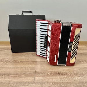Op de afbeelding: Een rode accordeon met witte en zwarte toetsen en een zwarte draagtas. De accordeon heeft een getextureerde afwerking en de merknaam "Corsuma" is zichtbaar. Het instrument staat op een houten vloer.