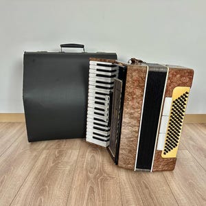 Op de afbeelding: Een bruin en beige accordeon met witte en zwarte toetsen, naast een zwarte draagtas. Het accordeon heeft een decoratief patroon op de voorkant en een geel en zwart rooster. De koffer heeft een handvat aan de bovenkant.