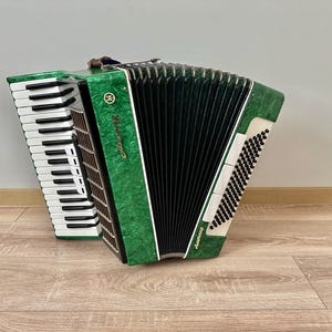 Op de afbeelding: Een groene en zwarte accordeon met witte toetsen en een zwarte balg. Op de accordeon staat het woord "Akkordeon". Het instrument staat op een houten vloer.