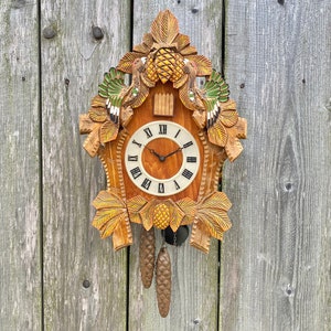 Può includere: Un orologio a cucù in legno con un design intagliato di uccelli e foglie. Il quadrante dell'orologio ha numeri romani e una lancetta nera. L'orologio è appeso a una parete di legno.