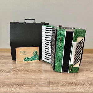 Op de afbeelding: Een groen en zwart accordeon met witte toetsen en een zwarte draagtas. Er is ook een kleine kaart met groene tekst en illustraties. Het accordeon staat op een houten vloer.