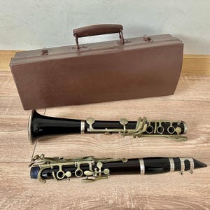 May include: A black clarinet with silver keys and a brown case with a handle. The clarinet is lying on a wooden surface. The case is brown and rectangular.