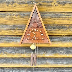 May include: A wooden cuckoo clock in a triangular shape, featuring Roman numerals and decorative carvings. The clock has a pendulum with pinecone weights and is mounted on a weathered wooden wall.