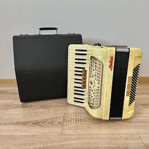 Op de afbeelding: Een wit en zwart accordeon met een bloemenpatroon en het woord "Hohner" erop gedrukt. Het accordeon staat op een houten vloer naast een zwarte draagtas.