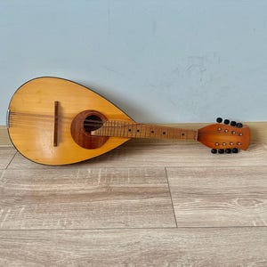 Peut inclure: Mandoline en bois avec un corps brun clair et un manche brun plus foncé. L'instrument possède des cordes, une rosace et des chevilles d'accord. Il repose sur une surface en bois, avec un mur bleu clair en arrière-plan.