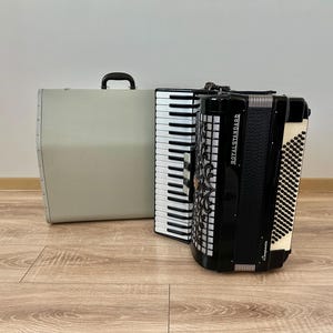 Op de afbeelding: Een zwart-wit Royal Standard accordeon met een toetsenbord en balg, naast een grijze draagtas. De accordeon heeft decoratieve accenten en staat op een houten vloer.