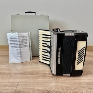 Op de afbeelding: Een zwart-wit Royal Standard accordeon met ivoorkleurige toetsen, een grijze koffer en bladmuziek. De accordeon heeft een zwarte balg en een decoratief ivoorkleurig paneel met ronde gaten. De koffer heeft een zwarte handgreep.