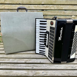 Op de afbeelding: Een zwart-witte accordeon met het woord "Monika" erop gedrukt. De accordeon staat op een houten bank met een grijze koffer ernaast.