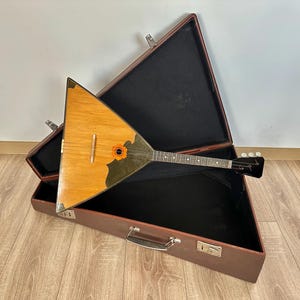 May include: A triangular balalaika with a light wood body, a dark green pickguard, and a floral soundhole rosette. The instrument features a long neck with frets and a black headstock, resting inside an open, brown, triangular hardshell case.