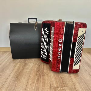 Op de afbeelding: Een rood en zwart accordeon met witte knoppen en het woord "Орфей" verticaal afgedrukt. Achter het accordeon staat een zwarte koffer met een handvat en een sleutelhanger. Het accordeon staat op een houten vloer.