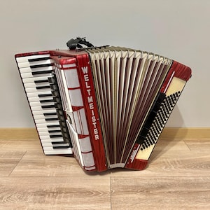 Pode incluir: Um acordeão vermelho e preto com a palavra "Weltmeister" impressa no lado. O acordeão tem um teclado branco e um fole preto.