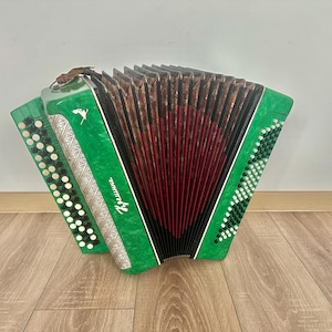 Könnte beinhalten: Ein grünes Akkordeon mit einem roten und braunen Balg. Das Instrument hat weiße und schwarze Knöpfe und silberne Verzierungen. Das Akkordeon steht auf einem Holzboden.
