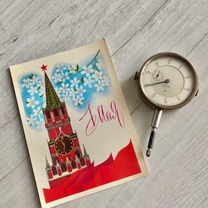 May include: A vintage postcard featuring a red clock tower with a white clock face and a red star on top. The tower is surrounded by white flowers and a red banner with the word "May" written in cursive. A silver dial gauge is also pictured.