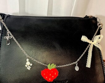May include: A black leather handbag with a silver chain strap and charms. The charms include a cross, a cluster of rhinestones, and a red embroidered strawberry. A white lace ribbon is attached to the strap.
