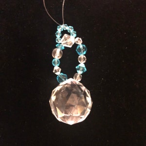 May include: A clear crystal ball pendant with a loop of blue and clear beads. The pendant hangs from a thin, clear string. The crystal ball is faceted, reflecting light. The beads are various shapes and sizes.
