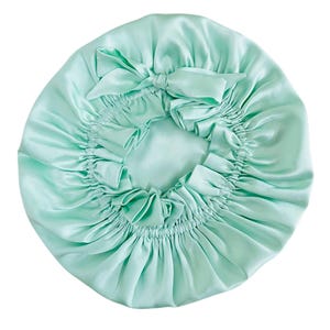 May include: A pale green satin bonnet with a gathered design and a bow on top. The bonnet is round and appears to be made of a soft, silky material. The bow is centered on the top of the bonnet.