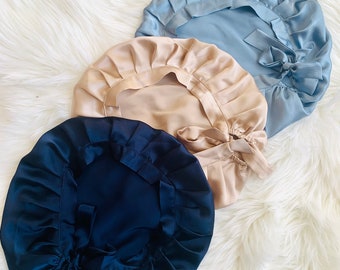 May include: Three satin hair bonnets in different colors: light blue, beige, and navy blue. Each bonnet has a gathered ruffle around the edge and a tie at the back.