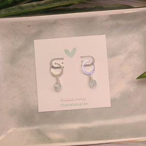 May include: A pair of silver hoop earrings with a light blue gemstone dangle. The earrings are displayed on a white card with the text "Handmade Jewelry Charmbangles".