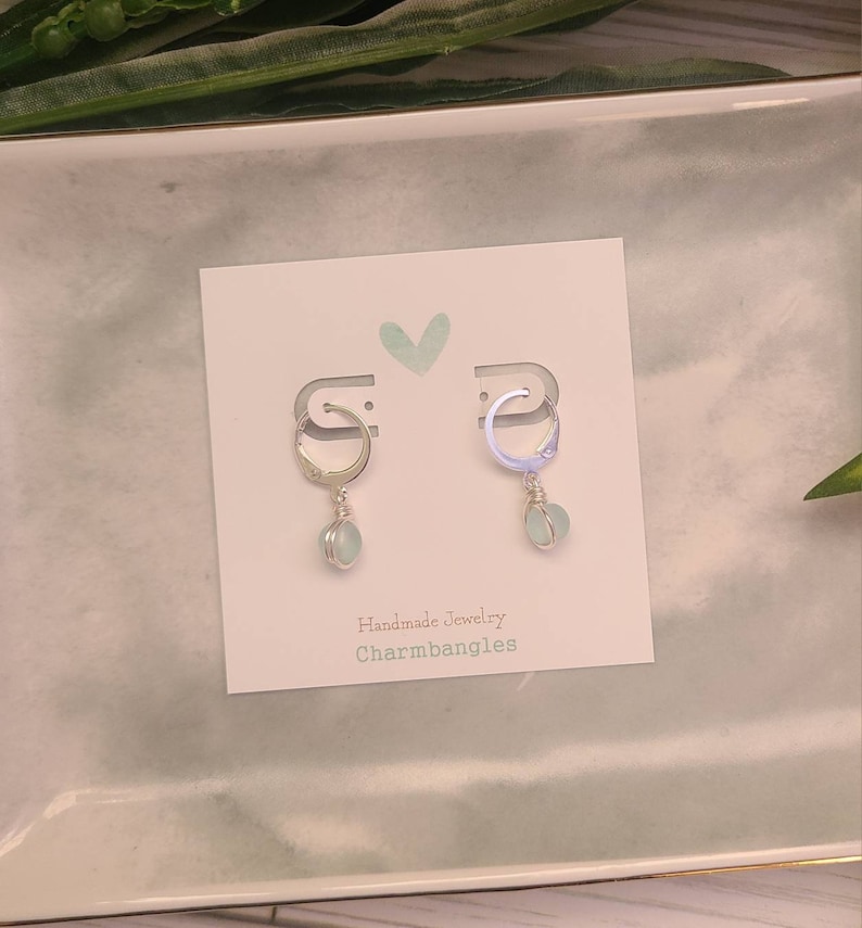 May include: A pair of silver hoop earrings with a light blue gemstone dangle. The earrings are displayed on a white card with the text "Handmade Jewelry Charmbangles".